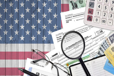 A graphic of the USA flag in the background and a pile of tax related documents, a magnifying glass, USA dollars, and a calculator